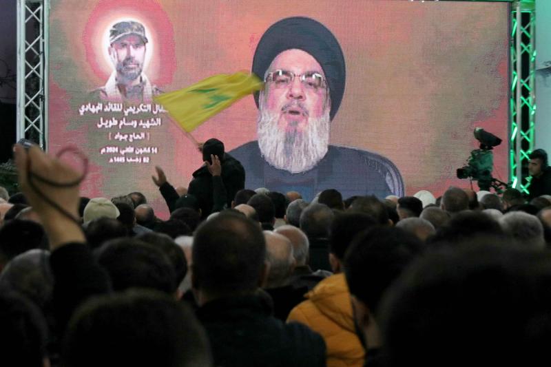 Güney Lübnan'ın Hirbet Selm beldesin Hizbullah Genel Sekreteri Hasan Nasrallah'ın konuşmasının yayınlandığı bir ekranın önünde Hizbullah bayrağı sallayan bir Hizbullah destekçisi, 14 Ocak 2024.jpeg