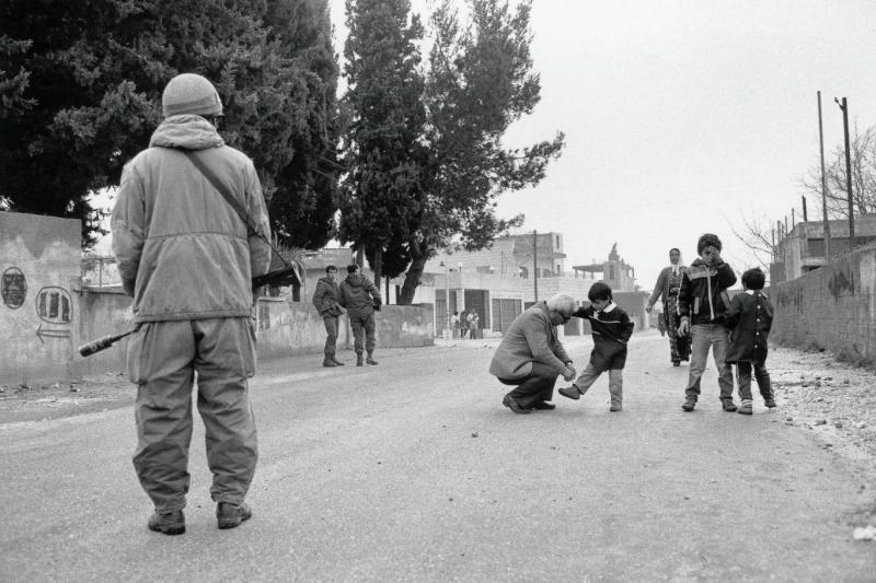Batı Şeria, 1980'lerin sonu: Birinci İntifada sırasında askeri ablukanın gölgesinde çocuğunun ayakkabısını bağlayan bir Filistinli
