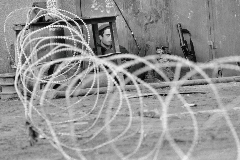 Birinci İntifada sırasında Gazze Şeridi'ndeki Nuseirat mülteci kampında barikatların arkasında bekleyen bir İsrail askeri. Bölgedeki yoğun güvenlik önlemlerini ve askeri ablukayı yansıtan bu an, dönemin tırmanan gerilimini gözler önüne seriyor (Ocak 1988)