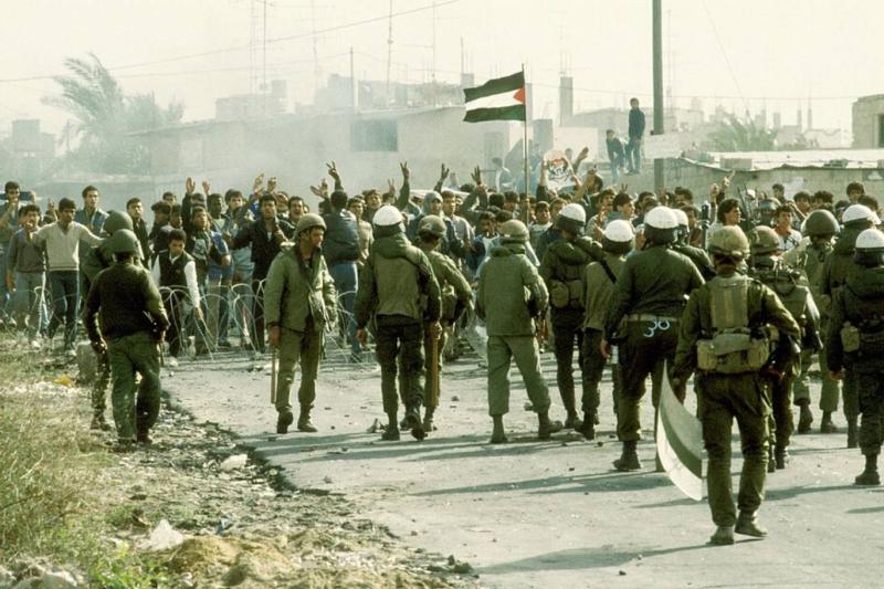 Gazze Şeridi, 1980'lerin sonu: Birinci İntifada sırasında tel örgülerle kapatılan sınır hattında, Filistin bayrağı açarak zafer işareti yapan göstericiler ile onlara müdahale eden çok sayıda İsrail askeri.