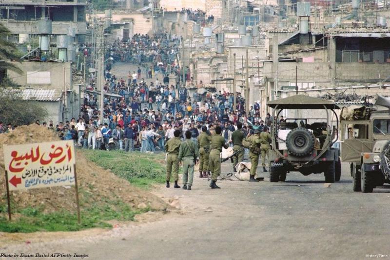 Batı Şeria, 1980'lerin sonu: Birinci İntifada sırasında bir Filistin mülteci kampında veya yerleşiminde, askeri araçlar eşliğinde yolu kapatan İsrail askerleri ve karşılarında toplanan kalabalık protestocu grup.