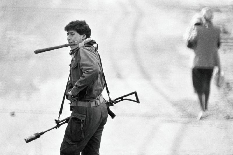 Birinci İntifada sırasında, ağzında askeri cop taşıyan ve sırtında tüfek bulunan bir İsrail askeri, bölgedeki Filistinli bir sivilin arkasından yürürken (1988)
